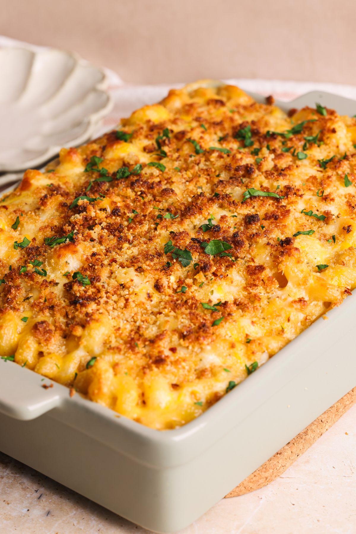 A rectangular baking dish filled with baked macaroni and cheese, topped with golden, crispy breadcrumbs and sprinkled with chopped parsley, sits on a countertop next to a stack of plates.