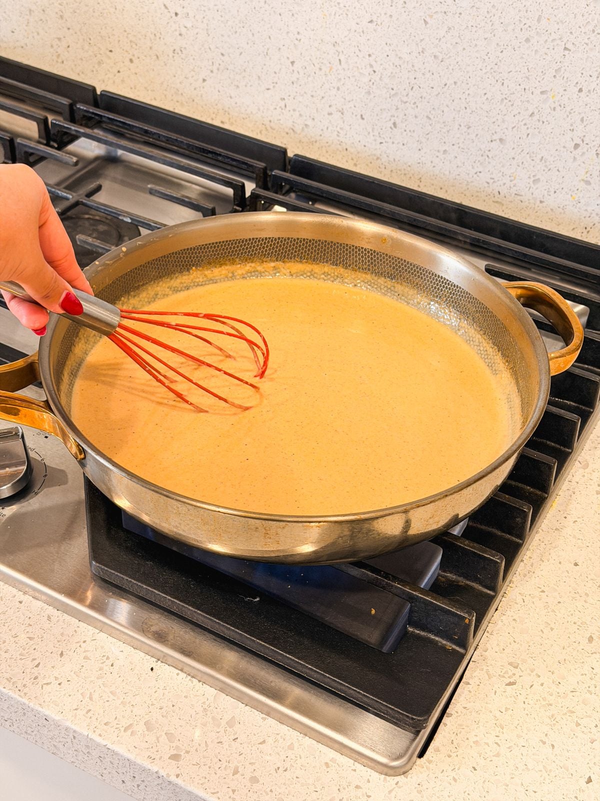A hand with red-painted nails is whisking a milk roux sauce in a large pan on a stovetop. The pan is on a gas burner, and the kitchen counter is light-colored.