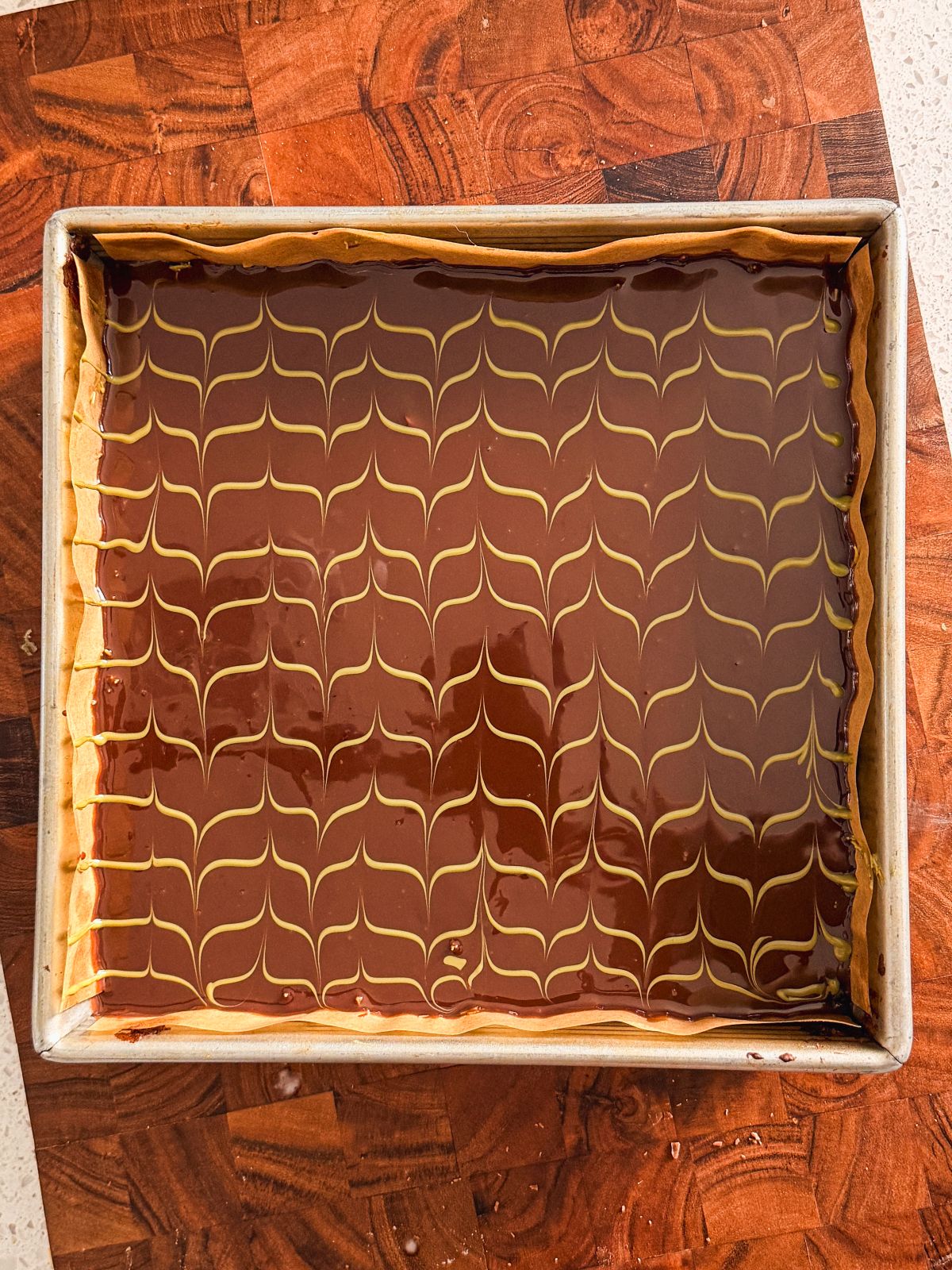 A rectangular tray of dessert with a glossy chocolate topping, decorated with a yellow wavy line pattern. The tray rests on a wooden surface.