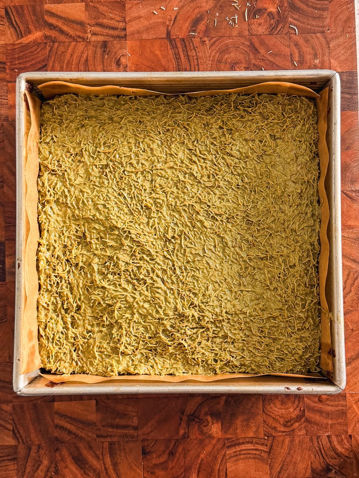 A square baking pan lined with parchment paper contains an even layer of pistachio knafeh on top of brownies on a wooden surface.