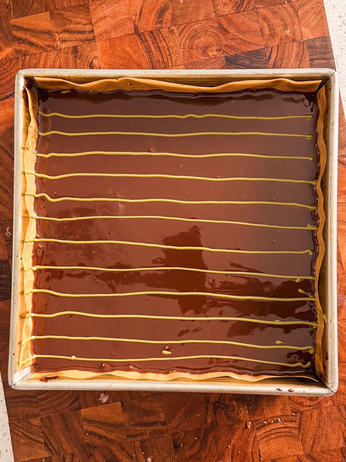 A square baking pan filled with brownies topped with a glossy ganache topping and evenly spaced thin green lines, set on a wooden surface.