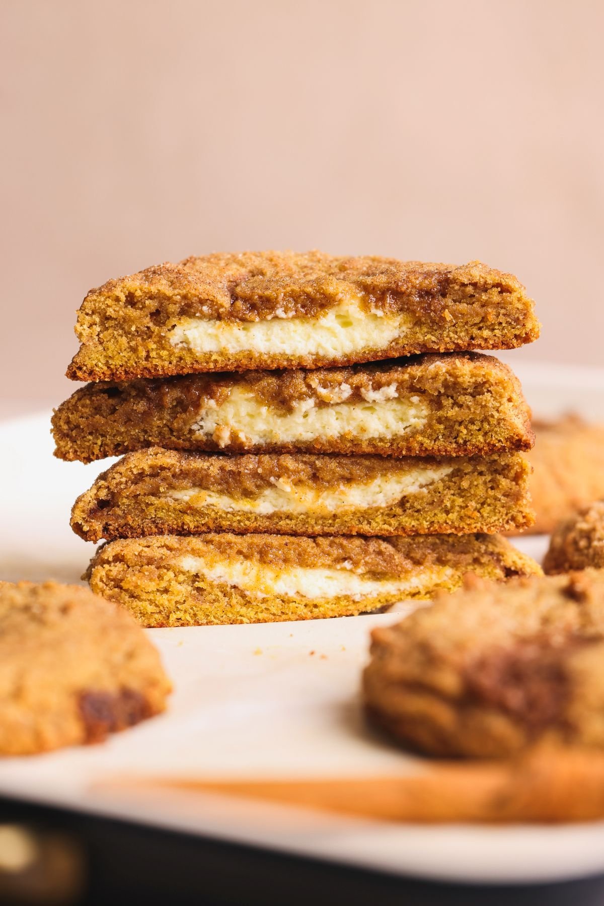 Stacked pumpkin cookies with creamy cheesecake filling are shown on a plate. One cookie is cut in half to reveal the soft filling inside, with more cookies blurred in the background.