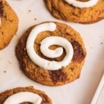 A close-up of a pumpkin cinnamon roll cookie topped with a swirl of cream cheese icing, resembling a cinnamon roll, on parchment paper with other similar cookies nearby.