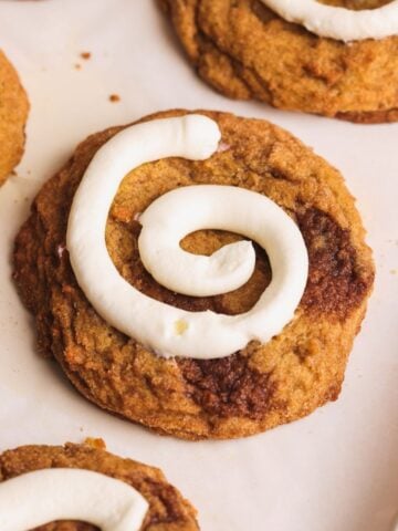 A close-up of a pumpkin cinnamon roll cookie topped with a swirl of cream cheese icing, resembling a cinnamon roll, on parchment paper with other similar cookies nearby.