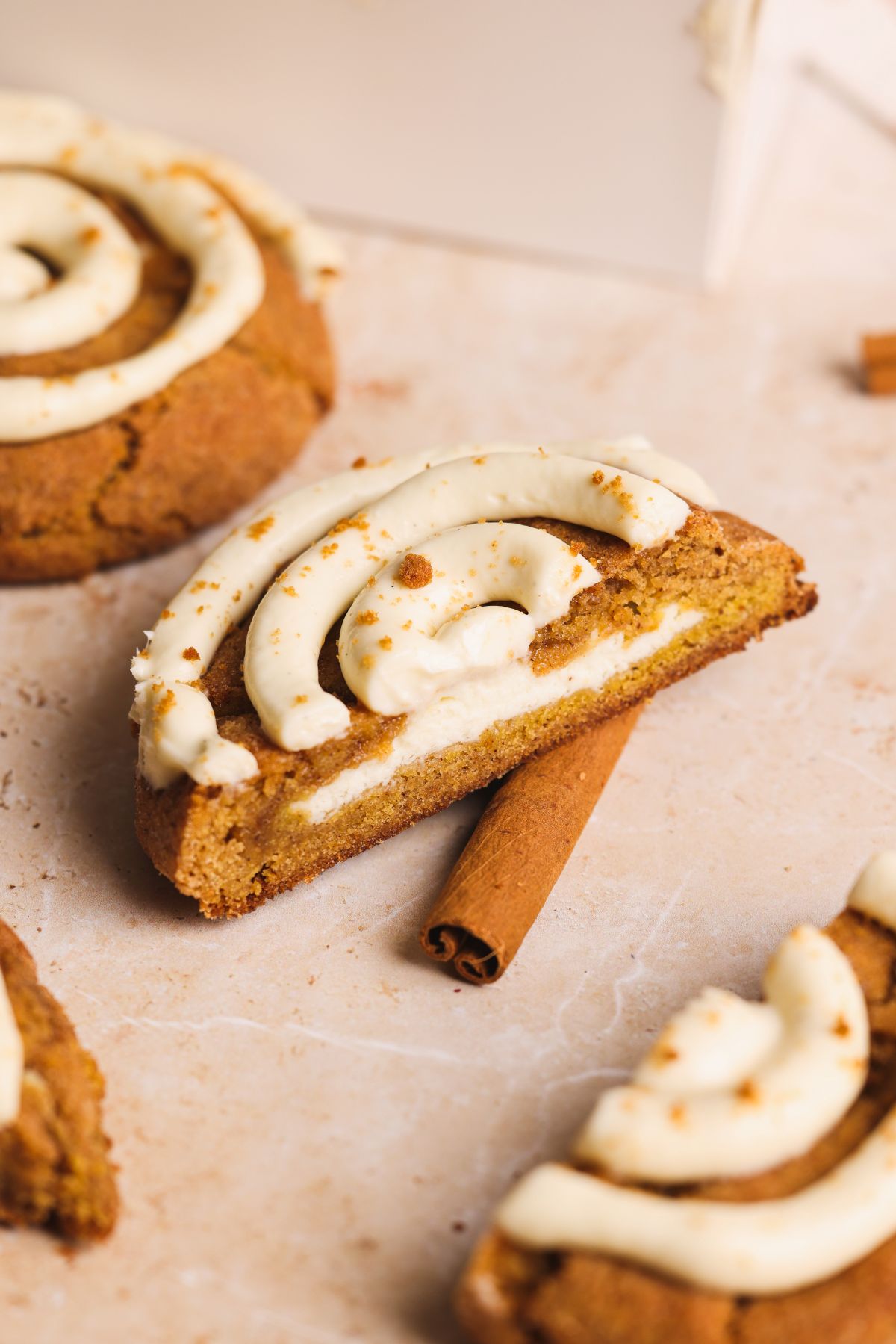 A thick pumpkin cinnamon roll cookie topped with swirled cream cheese icing sits on a cinnamon stick. Other similar treats are nearby on a light marble surface.