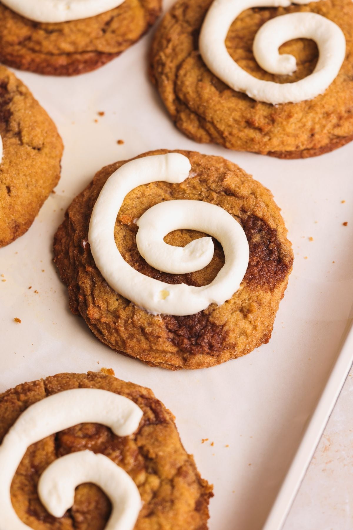 Close-up of freshly baked pumpkin cinnamon roll cookies with a spiral of cream cheese icing on top, arranged on a parchment-lined surface. The cookies have a golden-brown color and slightly crisp edges.