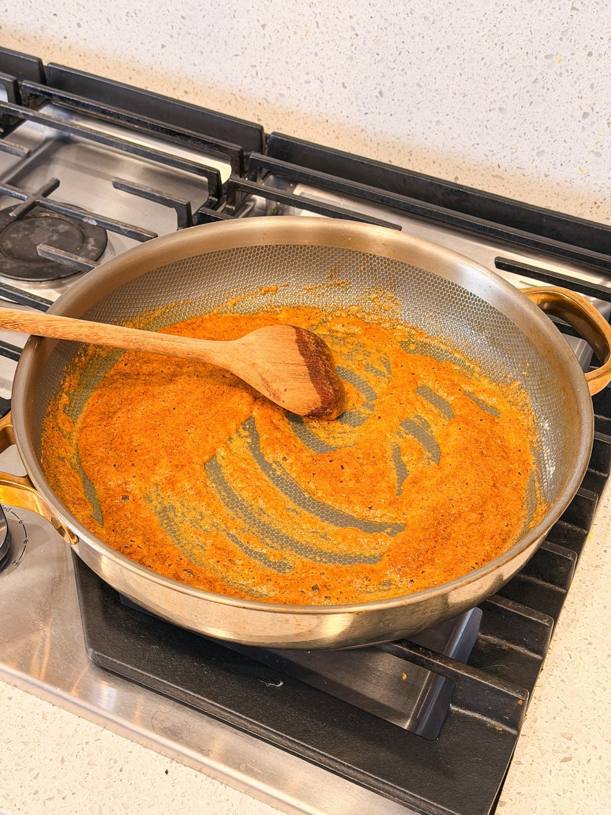 A stainless steel pan with spices in butter being stirred by a wooden spatula sits on a gas stove. The sauce appears partially cooked and spread along the bottom of the pan.