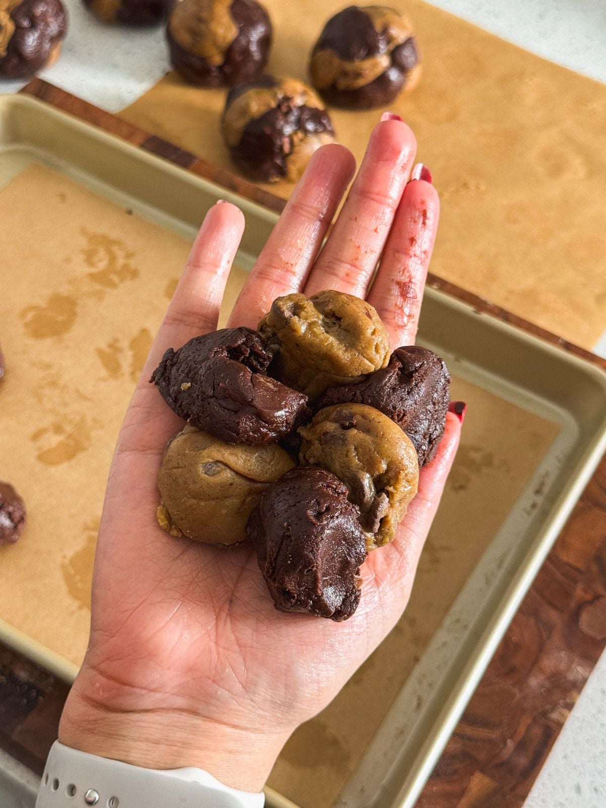 A hand holding six pieces of cookie dough, half chocolate and half plain, above a baking tray lined with parchment paper. More cookie dough pieces are on the tray in the background.