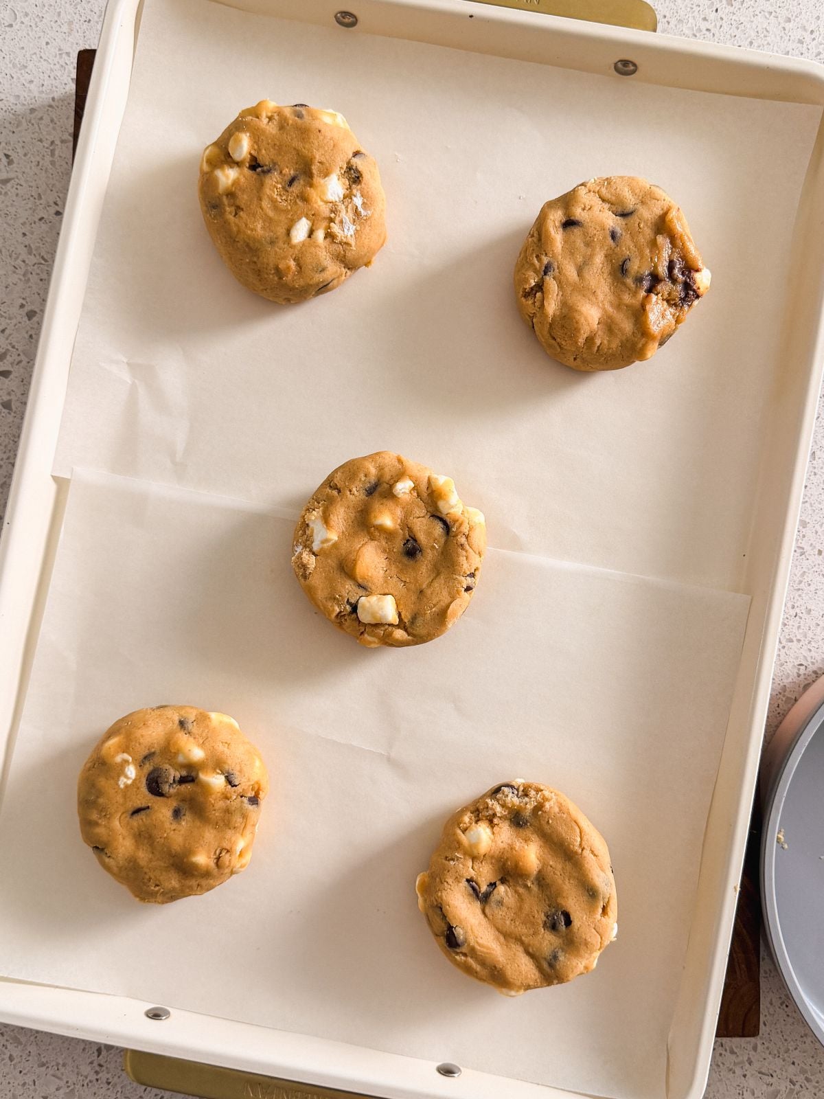 Five unbaked smores chocolate chip cookies are spaced out on a parchment-lined baking sheet, ready to be baked.