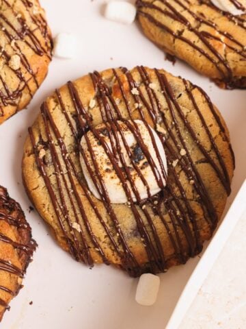 Close-up of a smores cookie topped with toasted marshmallows and drizzled with chocolate, placed on a light-colored tray. Crumbled graham crackers and small marshmallows are scattered nearby.