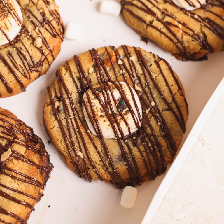 Close-up of a smores cookie topped with toasted marshmallows and drizzled with chocolate, placed on a light-colored tray. Crumbled graham crackers and small marshmallows are scattered nearby.
