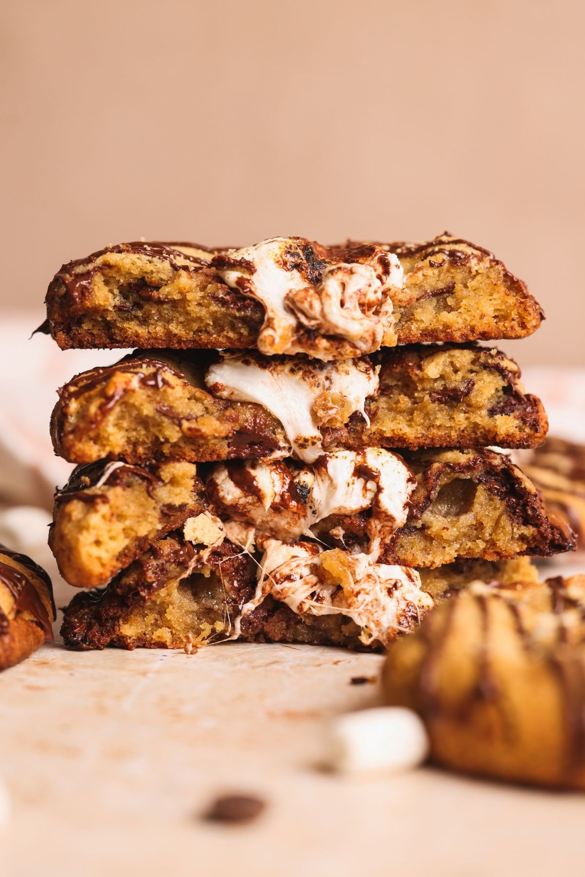A stack of gooey smores cookies with melted marshmallow oozing from the center, drizzled with chocolate and layered with bits of graham crackers, sits on a light surface against a blurred background.