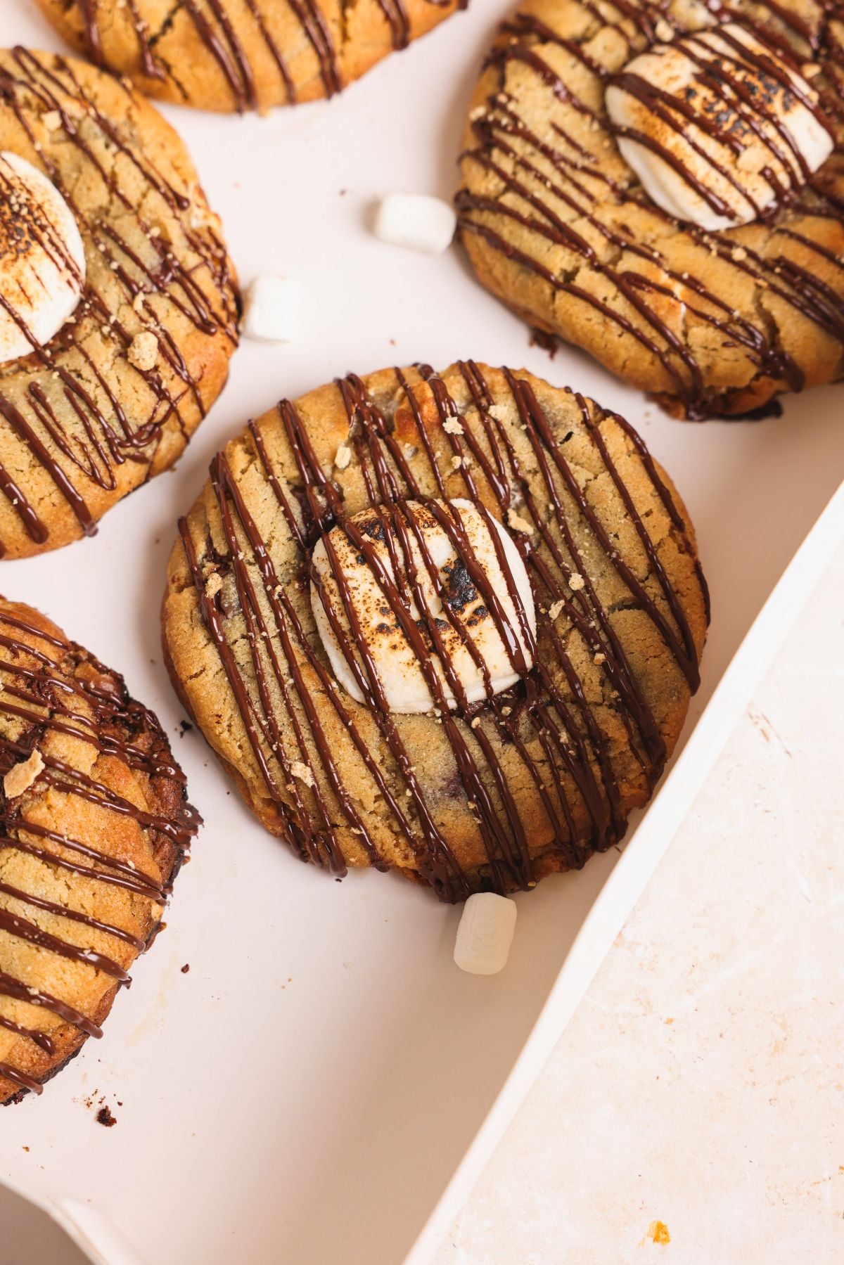 Smores cookies topped with toasted marshmallows and a drizzle of chocolate are arranged on a white tray, with graham crackers and mini marshmallows scattered around for an irresistible treat.