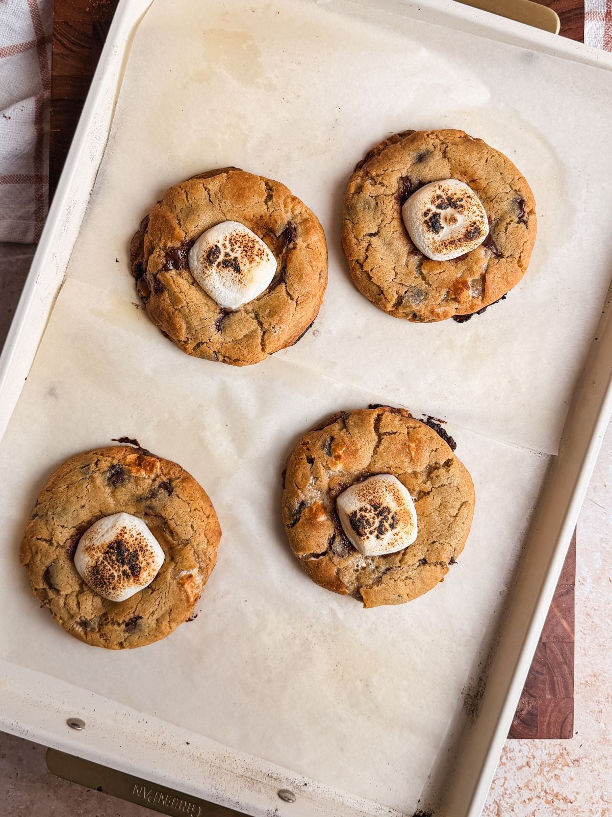 A baking sheet with four large smores cookies, each topped with a toasted marshmallow, on parchment paper. The cookies are golden-brown and slightly crispy at the edges.