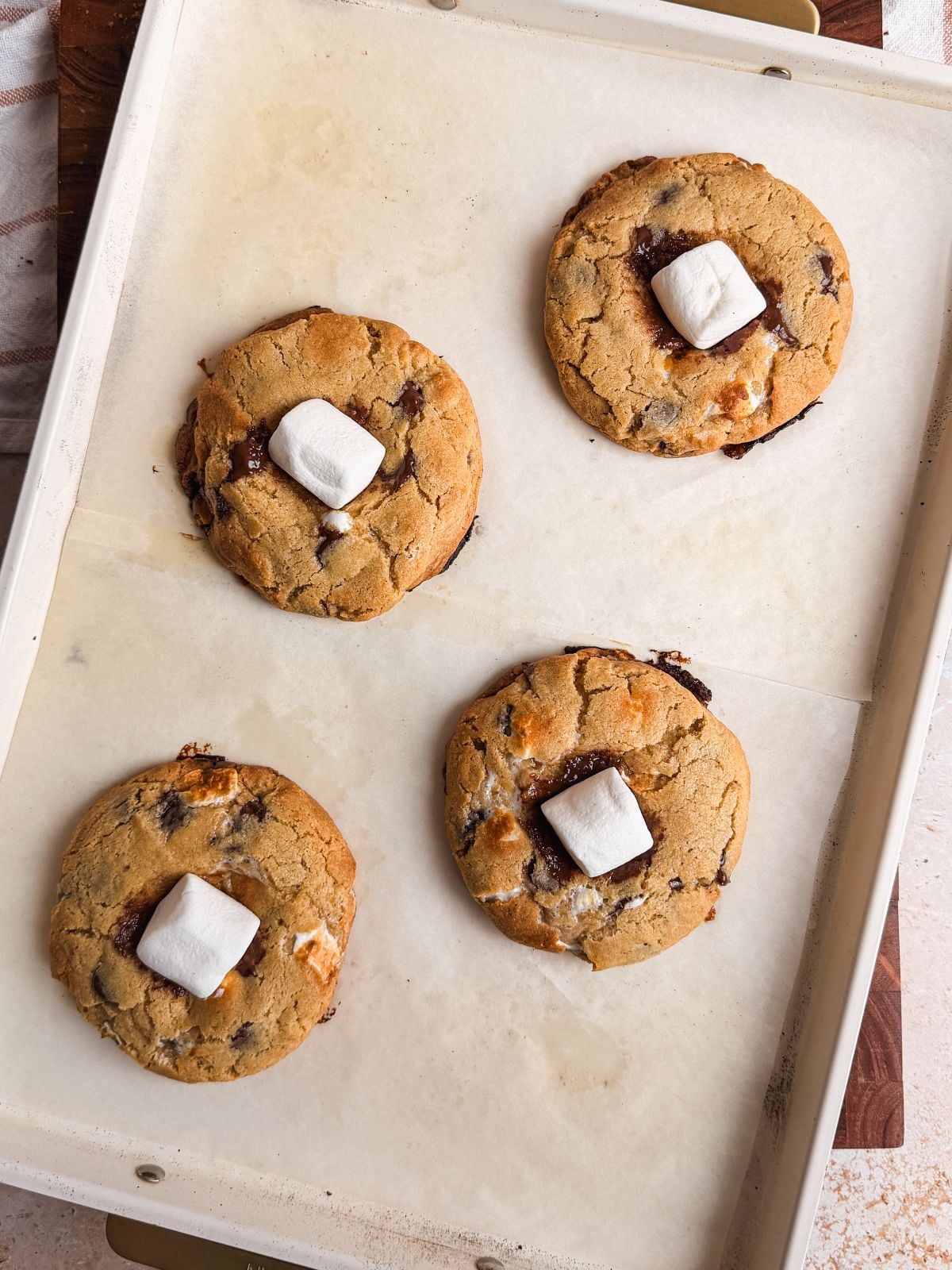 Four smores chocolate chip cookies topped with marshmallows are arranged on a parchment-lined baking sheet. The cookies are slightly golden with melty chocolate and marshmallow pieces visible.