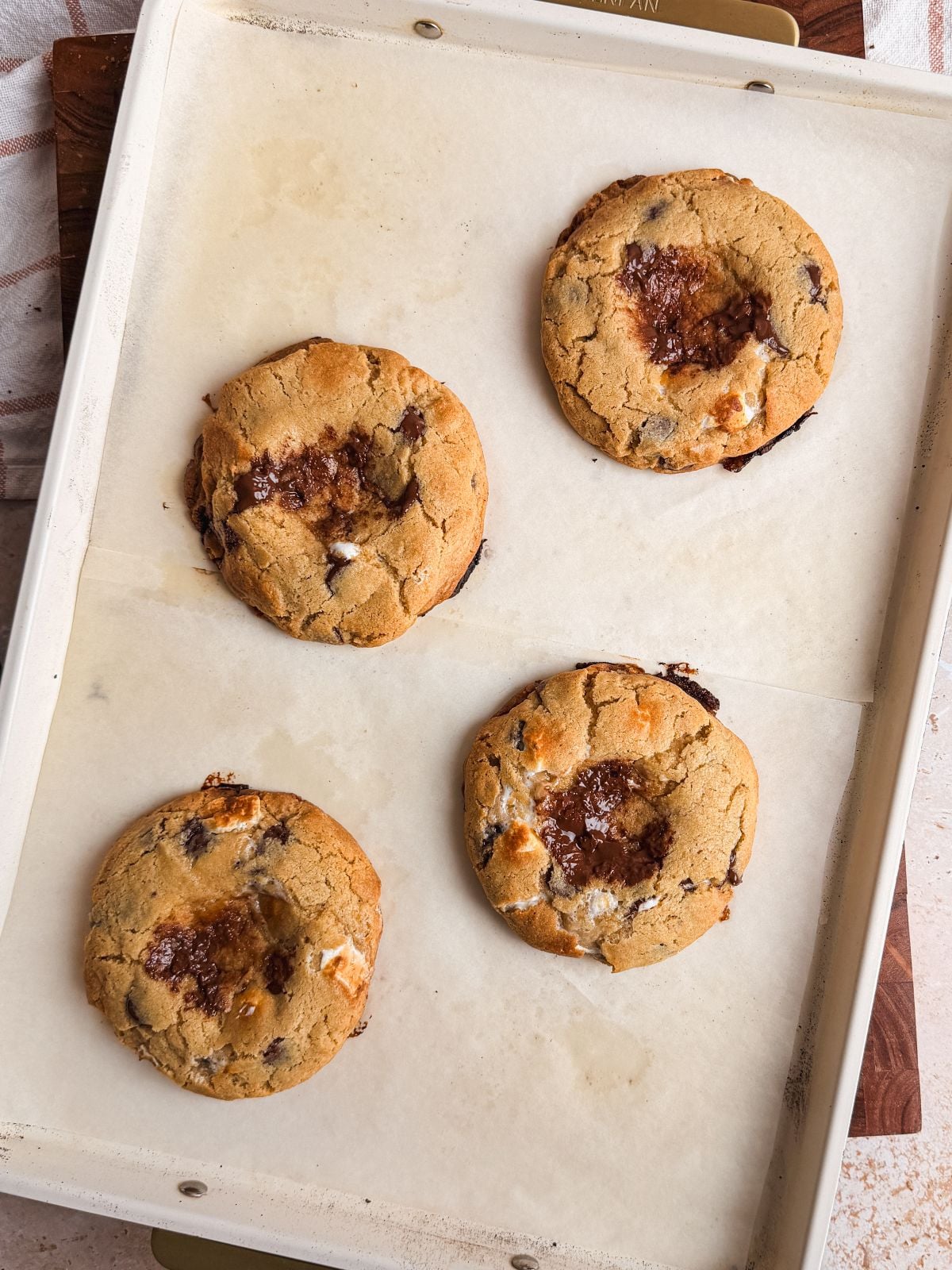 Four large smores cookies with pressed in centers are arranged on a parchment-lined baking sheet. The cookies appear freshly baked, with golden edges and gooey, caramelized centers.