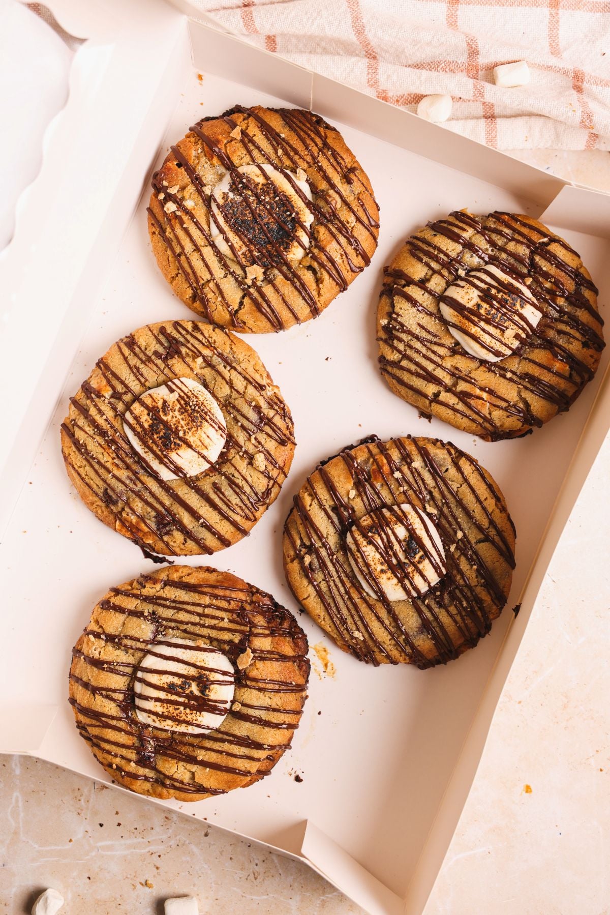 Five large smores cookies topped with toasted marshmallows and drizzled with chocolate are arranged in a white box on a light-colored surface. A patterned cloth is partially visible in the background.
