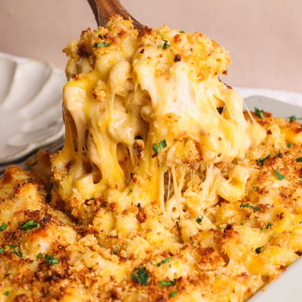 A close-up of baked macaroni and cheese being served, showing gooey melted cheese, golden-brown breadcrumbs, and a sprinkle of chopped parsley on top.