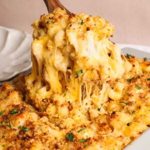 A close-up of baked macaroni and cheese being served, showing gooey melted cheese, golden-brown breadcrumbs, and a sprinkle of chopped parsley on top.