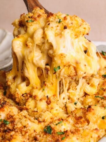 A close-up of baked macaroni and cheese being served, showing gooey melted cheese, golden-brown breadcrumbs, and a sprinkle of chopped parsley on top.