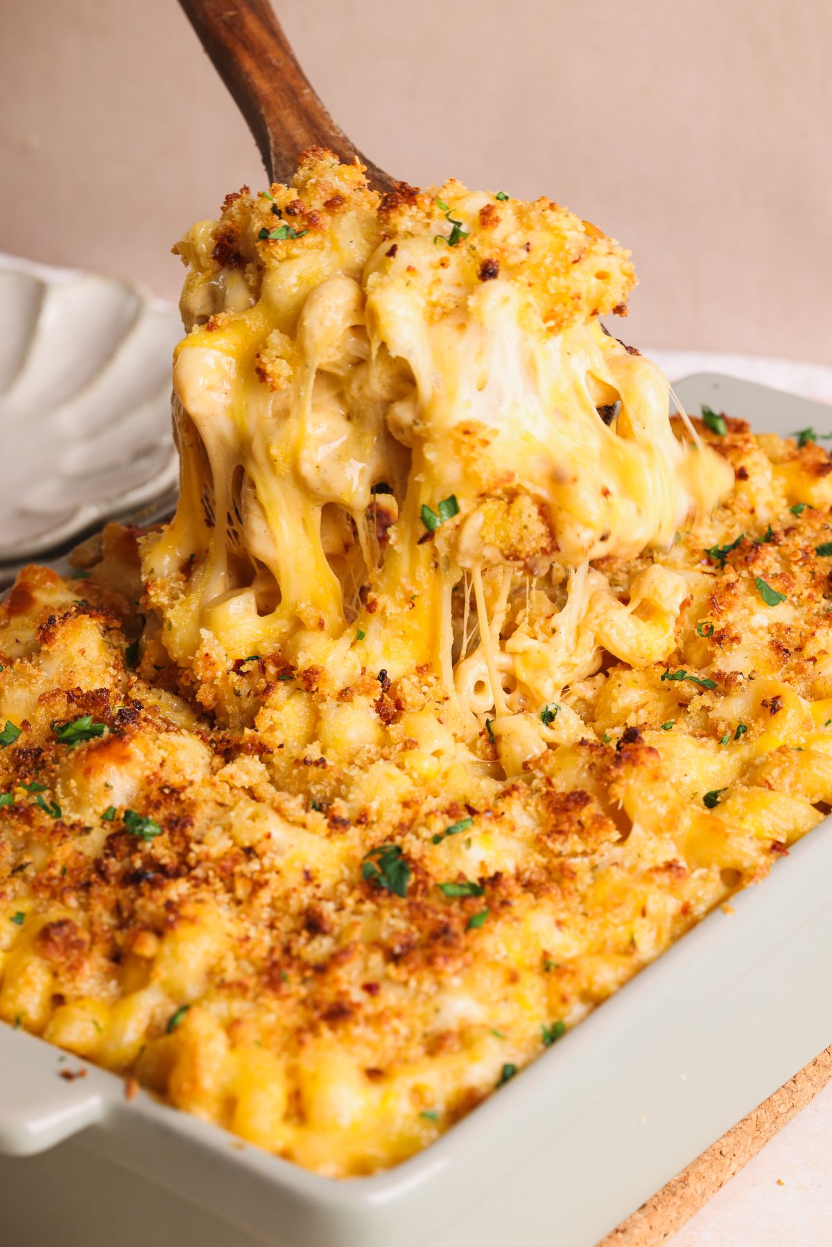 A wooden spoon lifts a serving of baked macaroni and cheese from a casserole dish, showing melted, stretchy cheese and a golden breadcrumb topping with chopped herbs.