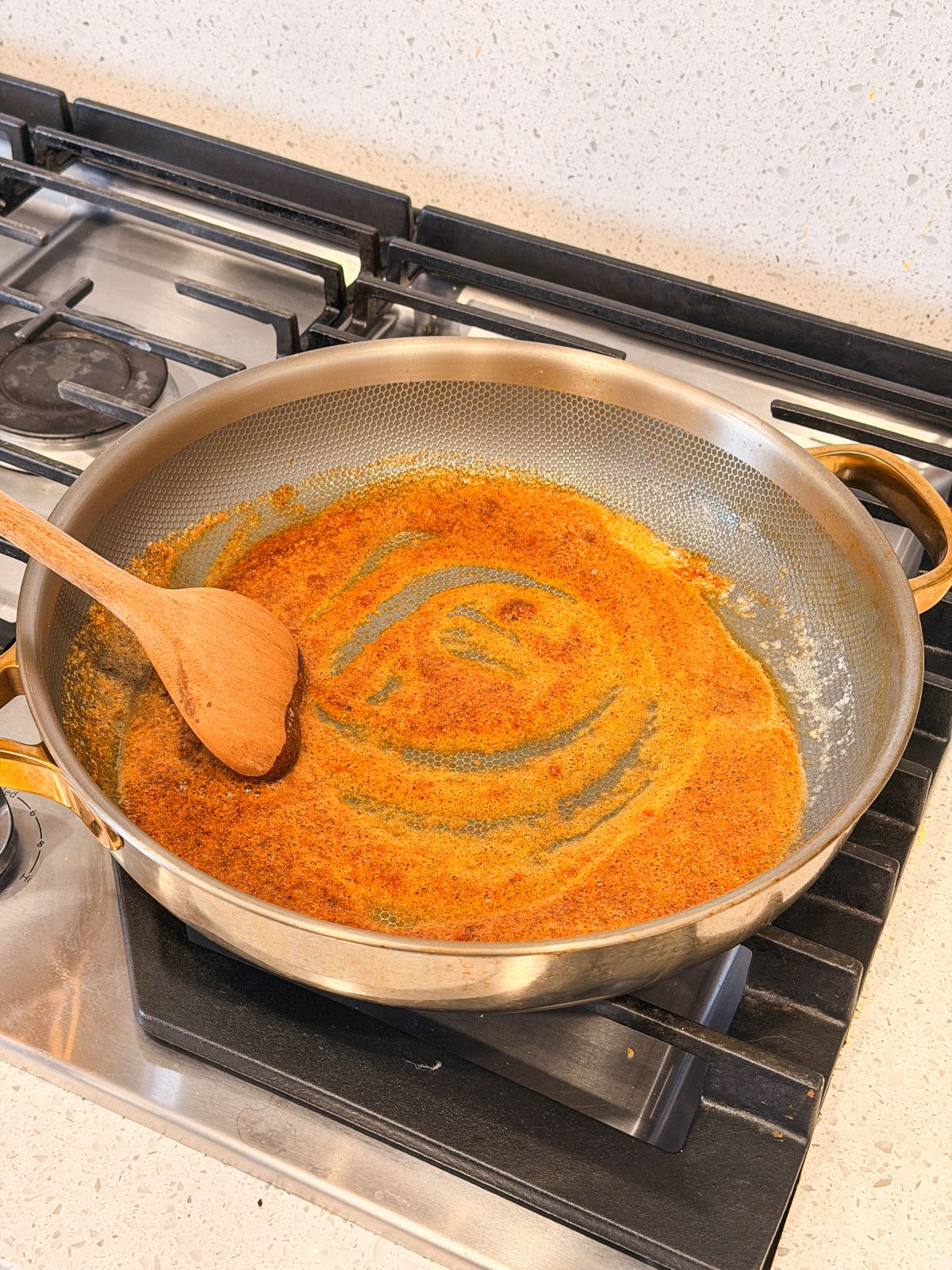 A wooden spoon rests in a large pan on a stove, stirring a golden-brown spice mixture or roux that is in the process of cooking.