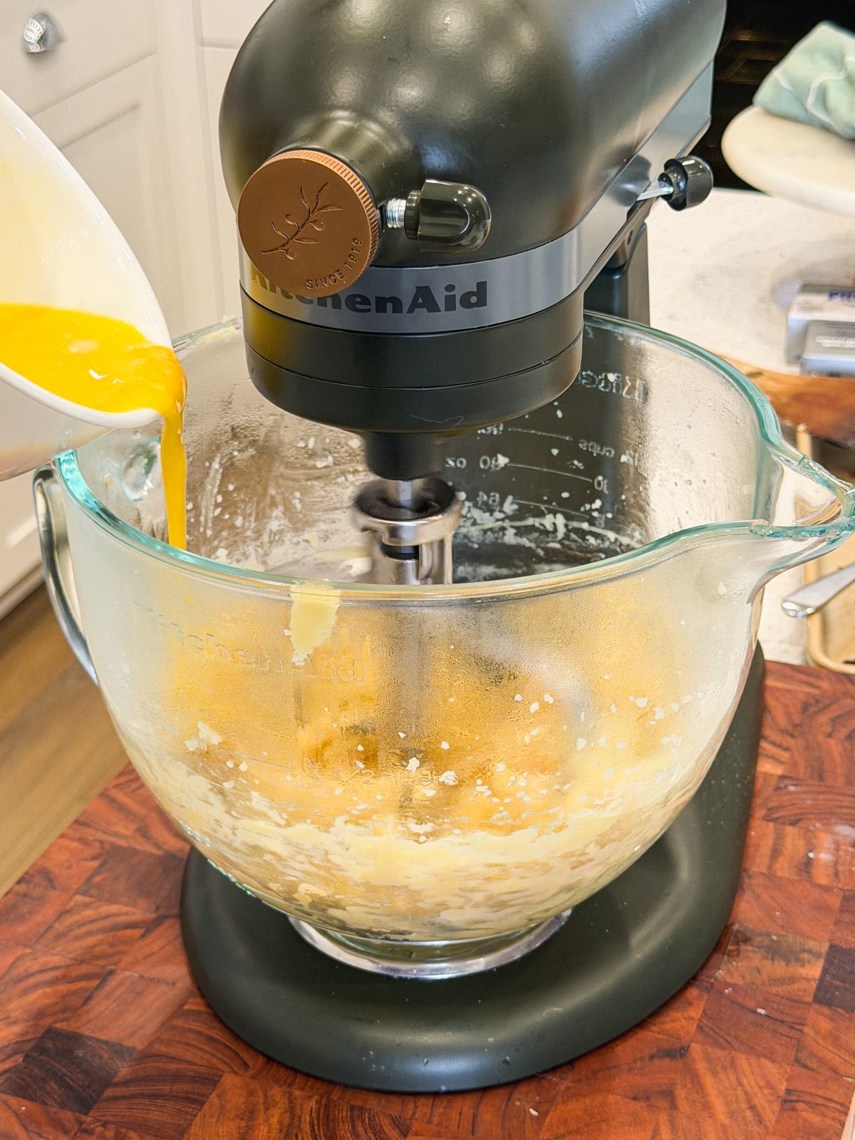 A close-up of a stand mixer blending choux pastry in a glass bowl while a person pours beaten eggs from a white bowl into the mixture. The mixer sits on a wooden counter in a kitchen.