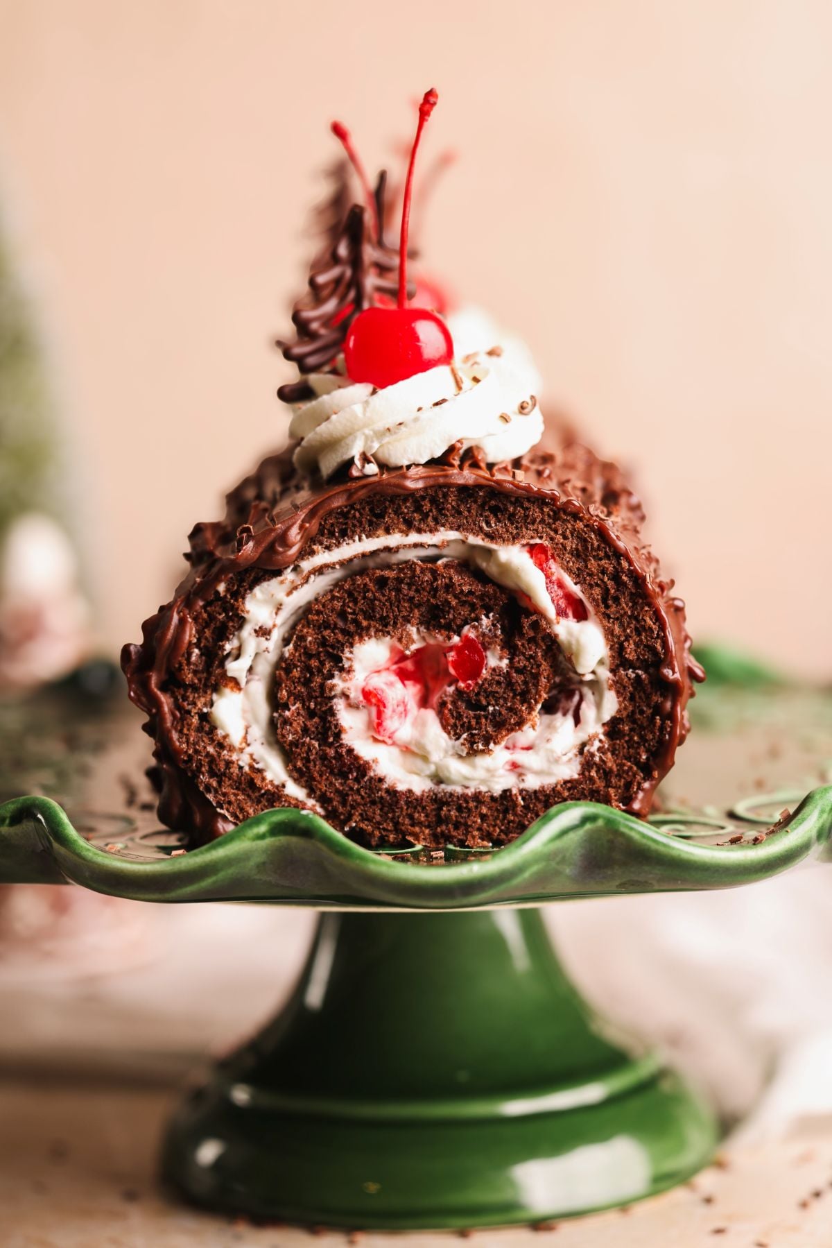 A close-up of a black forest chocolate Swiss roll cake filled with cream and cherries, topped with whipped cream, a cherry, and chocolate decoration, displayed on a green cake stand.