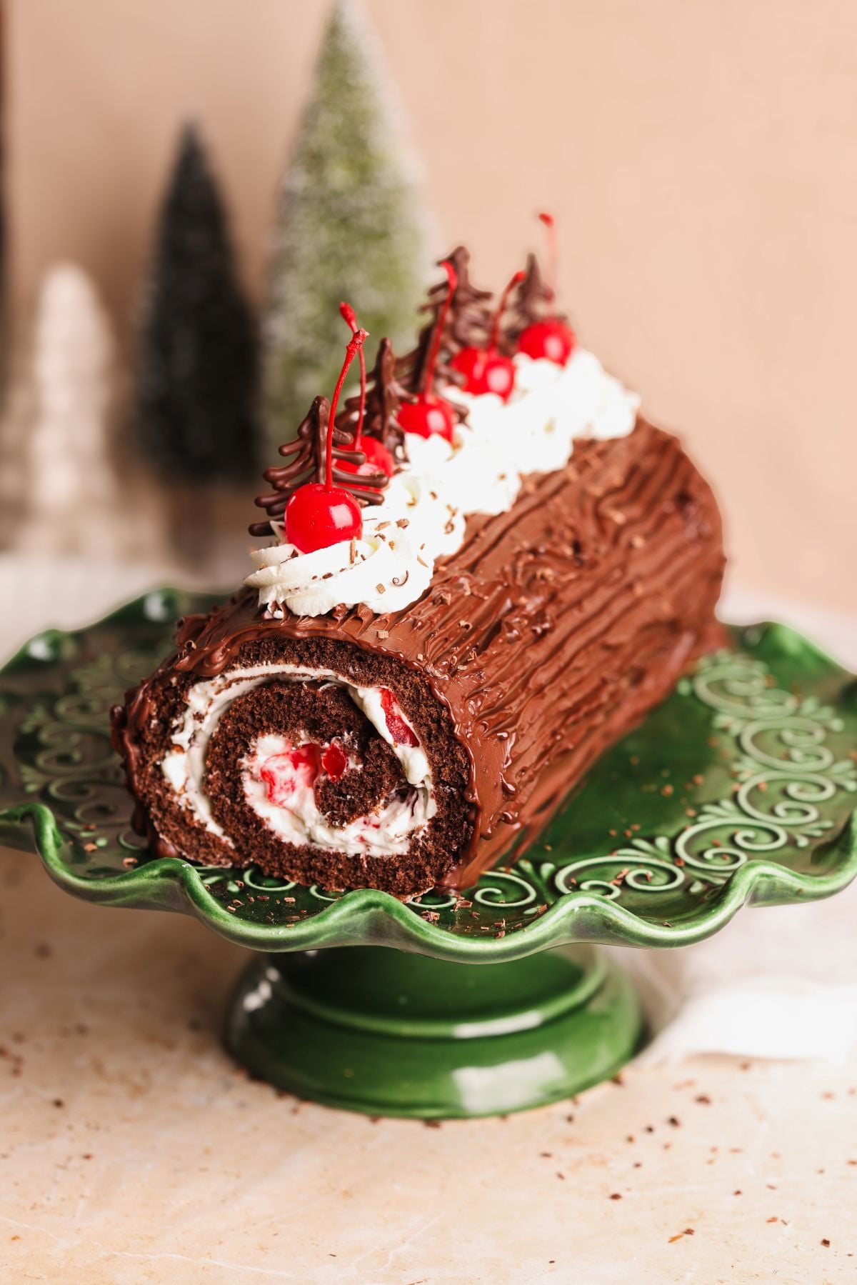 A black forest chocolate yule log cake topped with whipped cream, cherries, and chocolate decorations sits on a green cake stand, with blurred Christmas trees in the background.