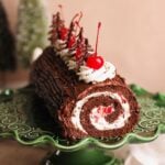 A black forest yule log cake with a cream and cherry filling, topped with whipped cream, cherries, and chocolate decorations, sits on a decorative green cake stand.