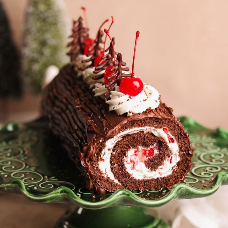 A black forest yule log cake with a cream and cherry filling, topped with whipped cream, cherries, and chocolate decorations, sits on a decorative green cake stand.