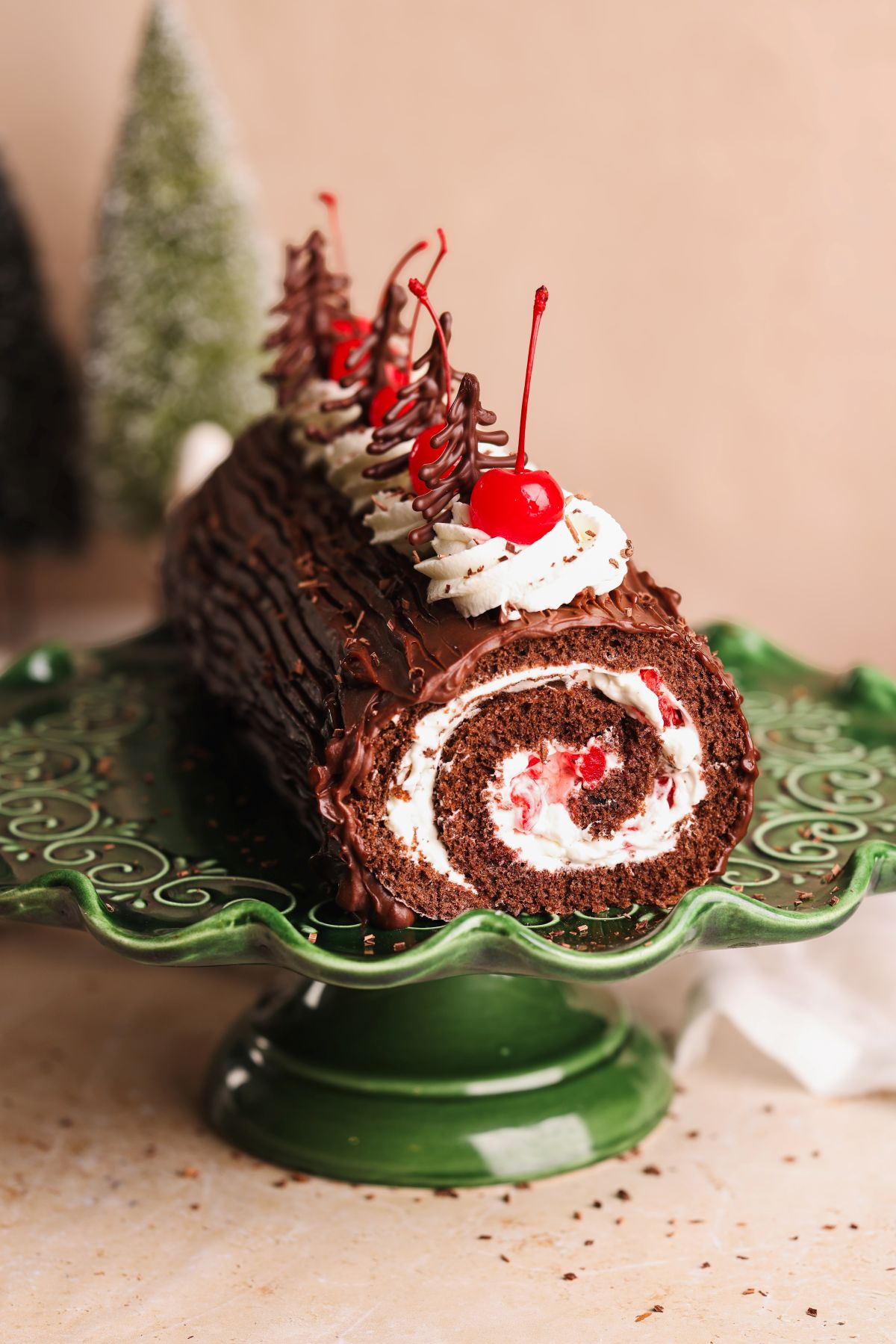 A chocolate yule log cake with cream and cherries inside, topped with whipped cream, cherries, and chocolate decorations, displayed on a decorative green cake stand.