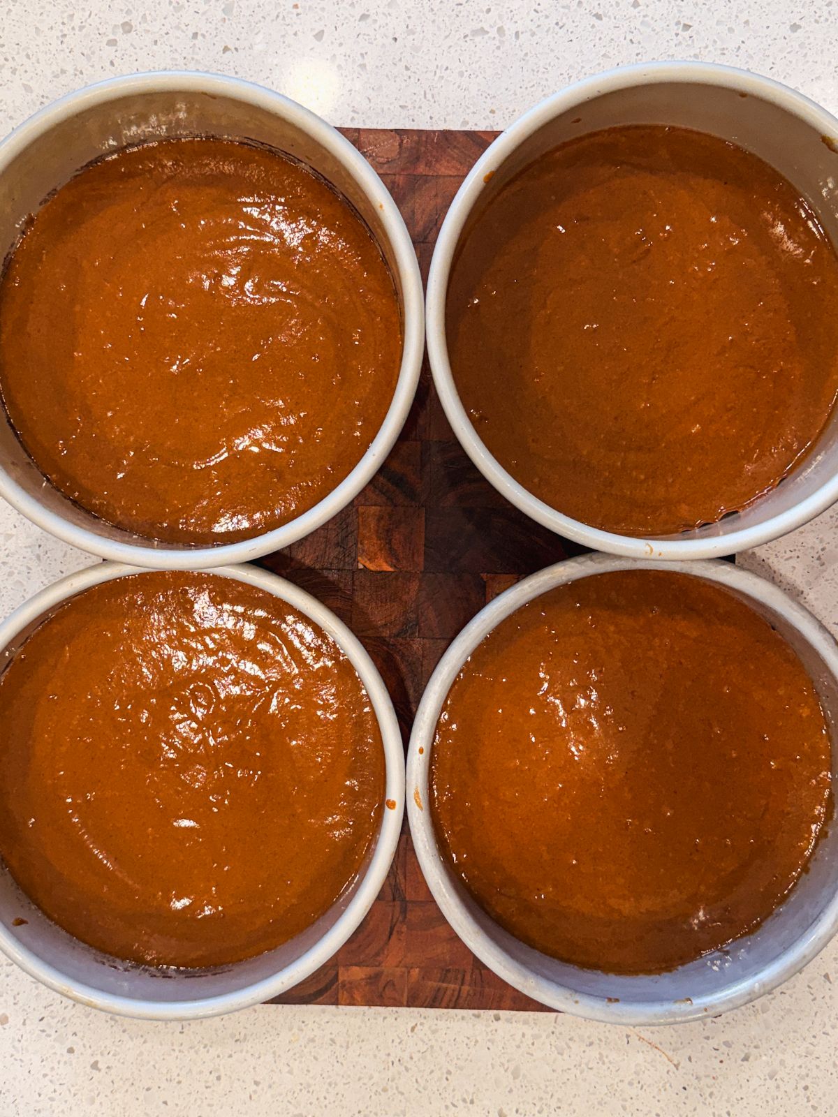Four round baking pans filled with smooth, gingerbread cake batter are arranged in a square on a wooden surface, ready to be baked. The countertop is light-colored and speckled.