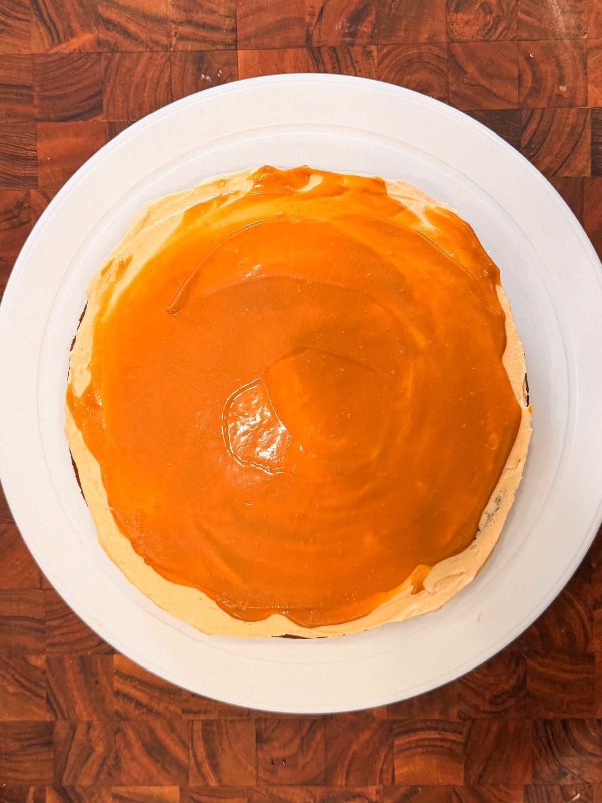 A round cake with frosting and dulce de leche cream on top sits on a white plate, placed on a wooden surface with a checkered pattern.
