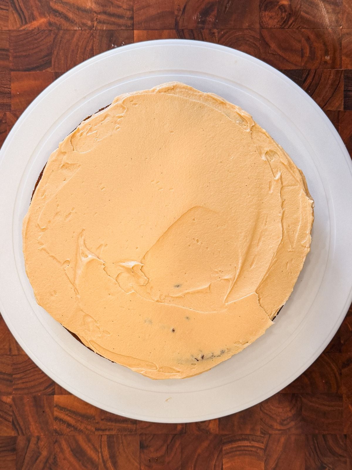 A round cake with smooth light orange frosting sits on a white plate atop a wooden surface. The cake appears homemade, with a slightly uneven layer of frosting on top.
