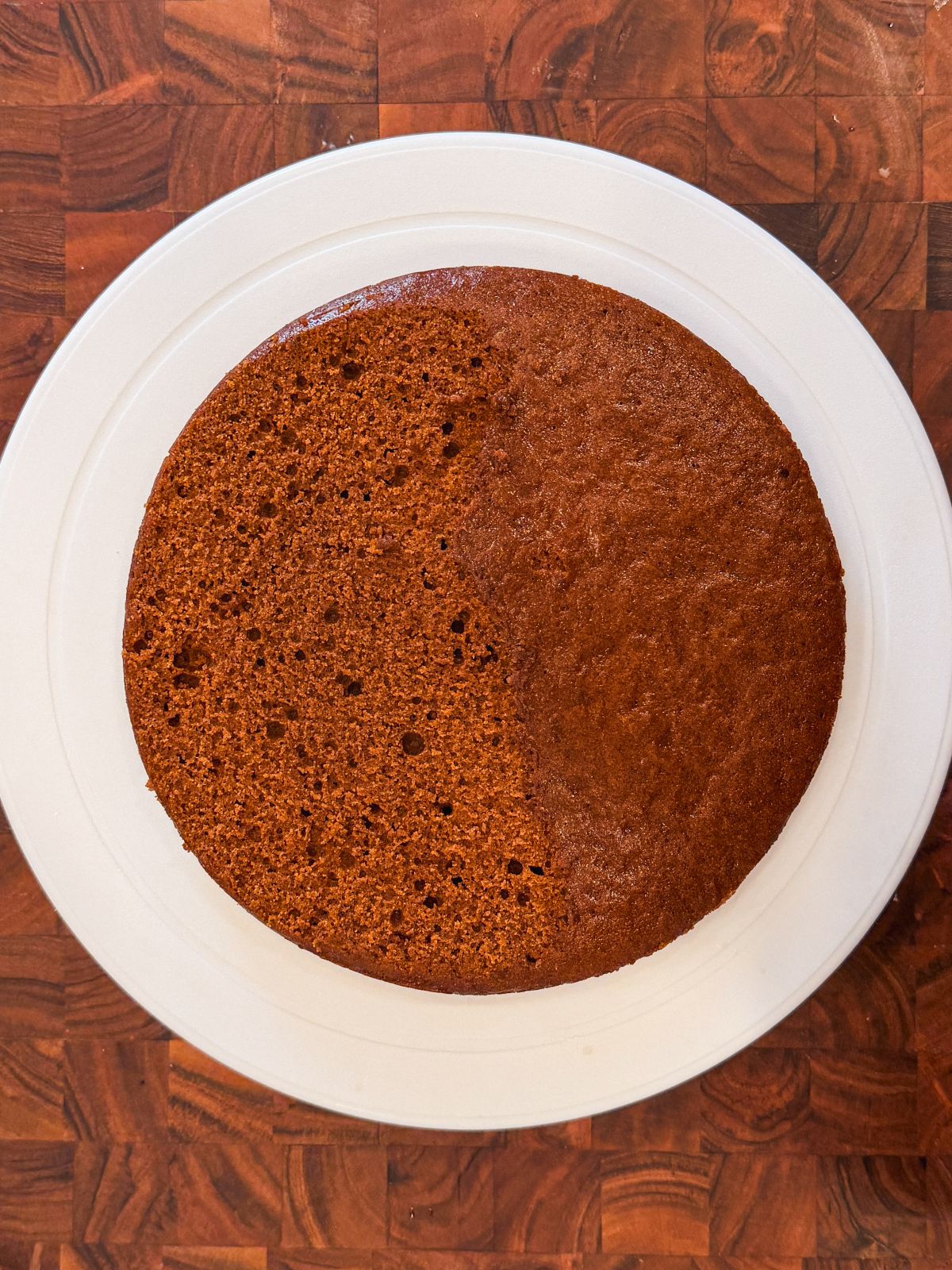 A round, brown gingerbread cake sits on a white plate atop a wooden surface. Half of the cakes top has been sliced off, revealing its airy, textured interior.