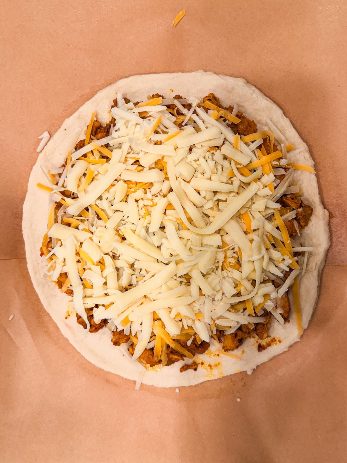 A raw pizza with shredded cheese and seasoned meat on a round dough base, placed on brown parchment paper.