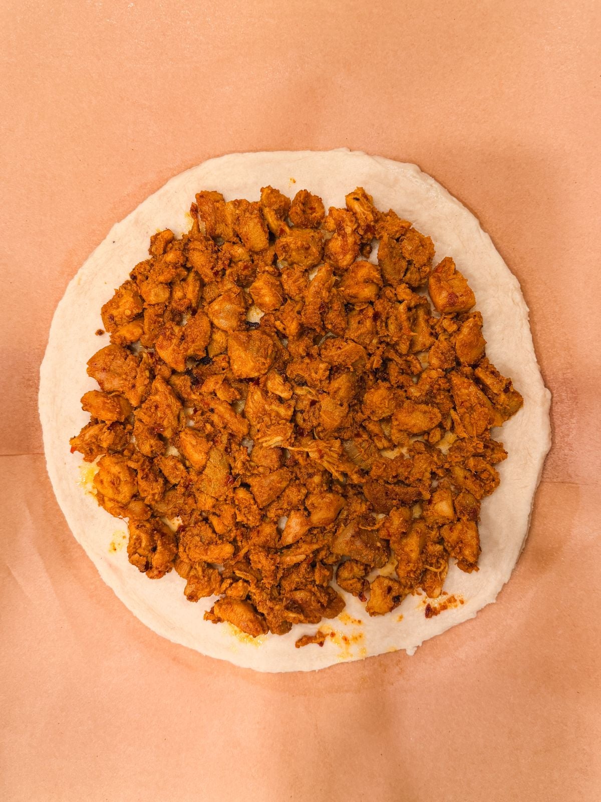 A round flatbread topped with a generous layer of seasoned, cooked chicken pieces, placed on a sheet of brown parchment paper.