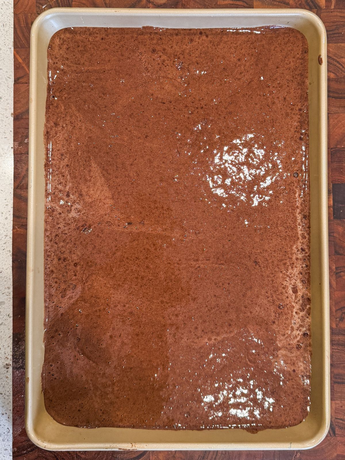 A baking sheet filled with an even layer of uncooked chocolate genoise cake batter sits on a wooden surface, ready to be baked.