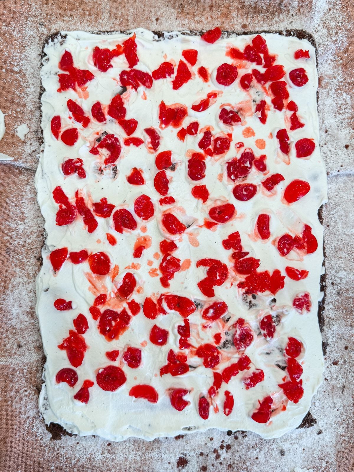 A rectangular sheet of chocolate cake topped with a layer of white cream and scattered with pieces of bright red cherries, sitting on a floured surface.