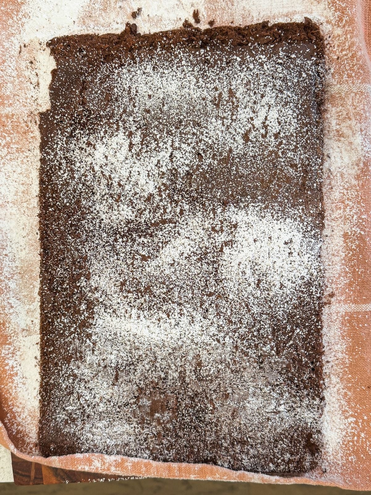 A large rectangular chocolate genoise cake rests on an orange kitchen towel, lightly dusted with powdered sugar. The cake appears to have been baked in a sheet pan