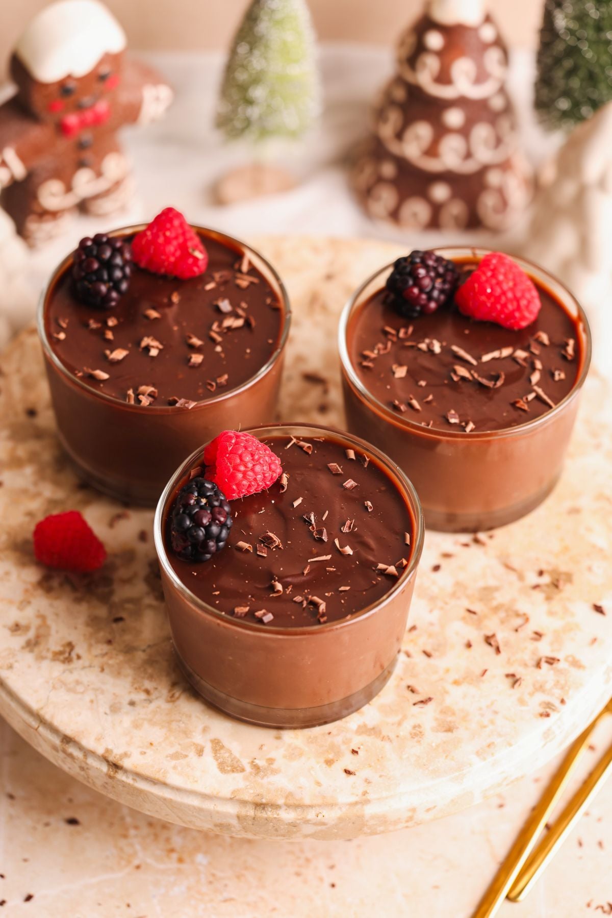 Three glass cups filled with chocolate pudding, topped with chocolate shavings, raspberries, and blackberries. They are placed on a marble surface, with festive decorations in the background.