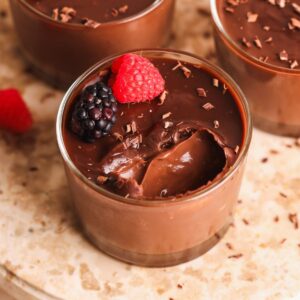 A glass cup filled with creamy chocolate pudding, topped with a raspberry, a blackberry, and chocolate shavings, sits on a marble surface. A spoonful of pudding has been taken out.