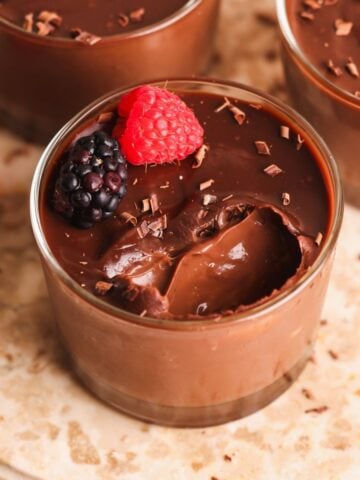 A glass cup filled with creamy chocolate pudding, topped with a raspberry, a blackberry, and chocolate shavings, sits on a marble surface. A spoonful of pudding has been taken out.