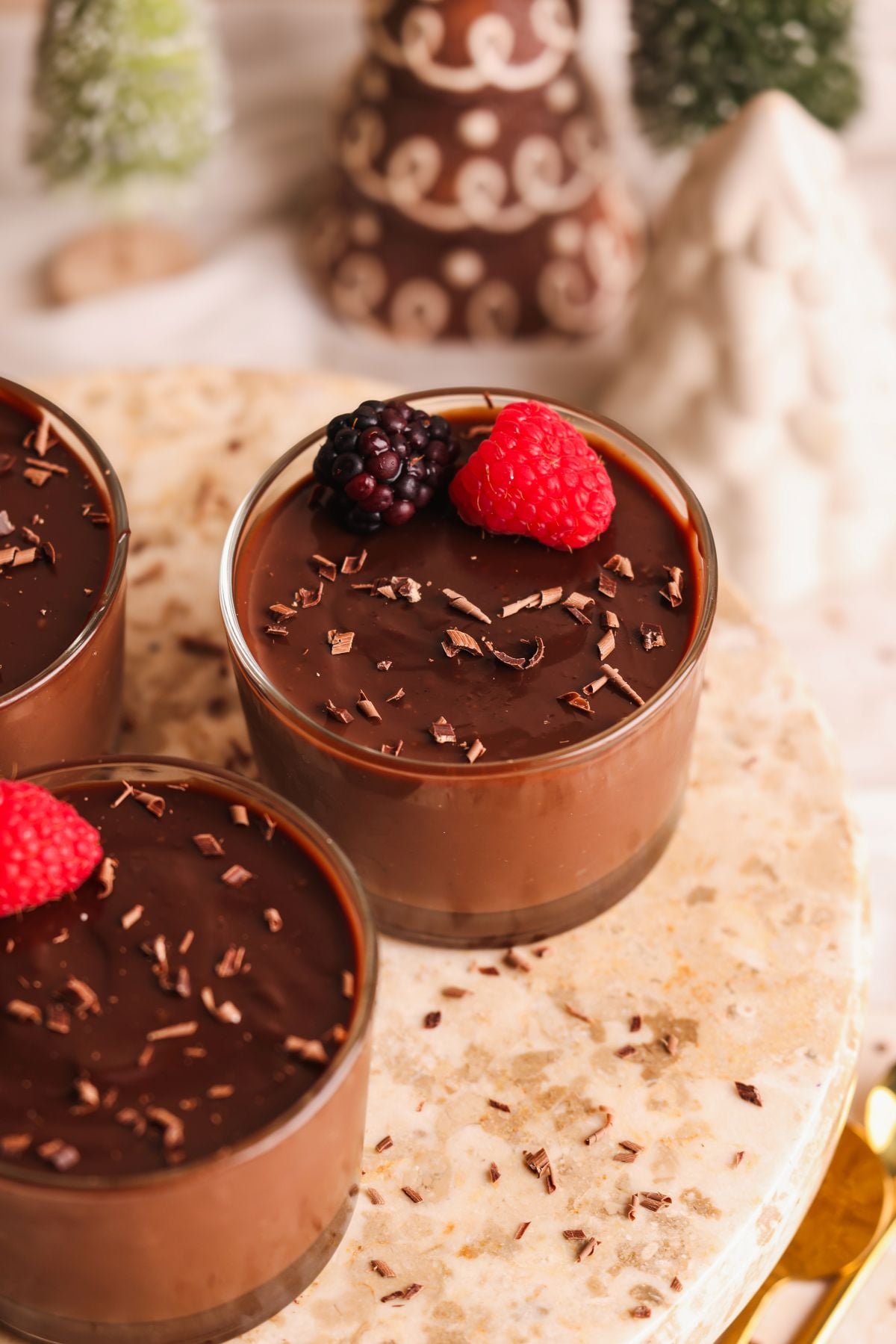 Three glass cups filled with chocolate pudding, topped with chocolate shavings and fresh blackberries and raspberries, are displayed on a marble surface. Blurred decorative trees are visible in the background.