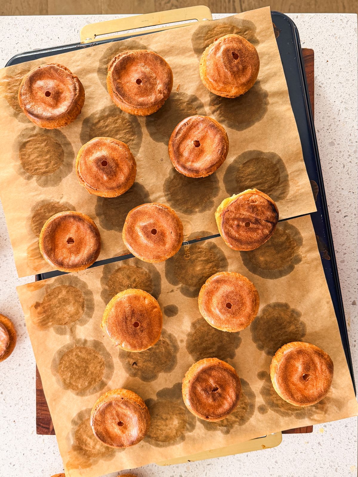 Golden brown choux pastry shells with small holes in the center are arranged in neat rows on parchment paper atop baking trays, casting soft, round shadows from the overhead light.