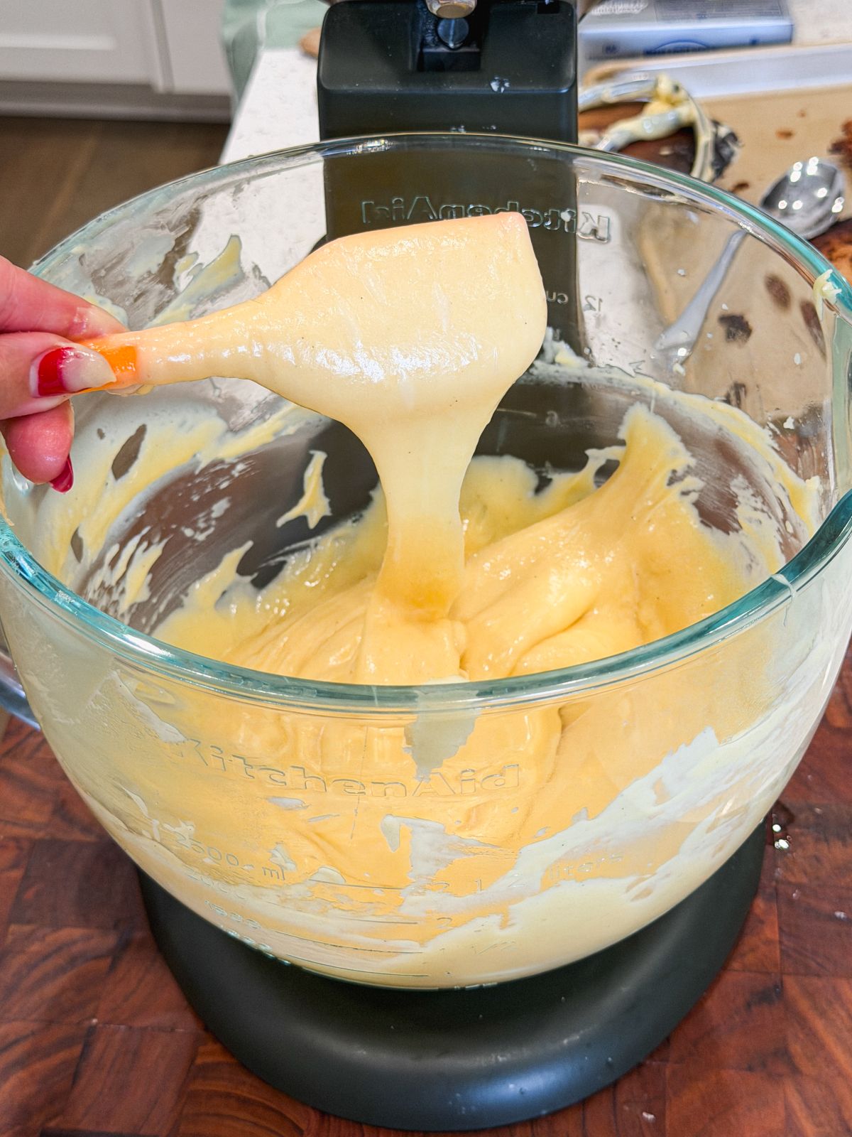 A hand lifts a thick, smooth, choux pastry from a glass mixing bowl attached to a stand mixer. The batter stretches as it is lifted, showing its glossy texture. The kitchen counter is messy with baking tools in the background.