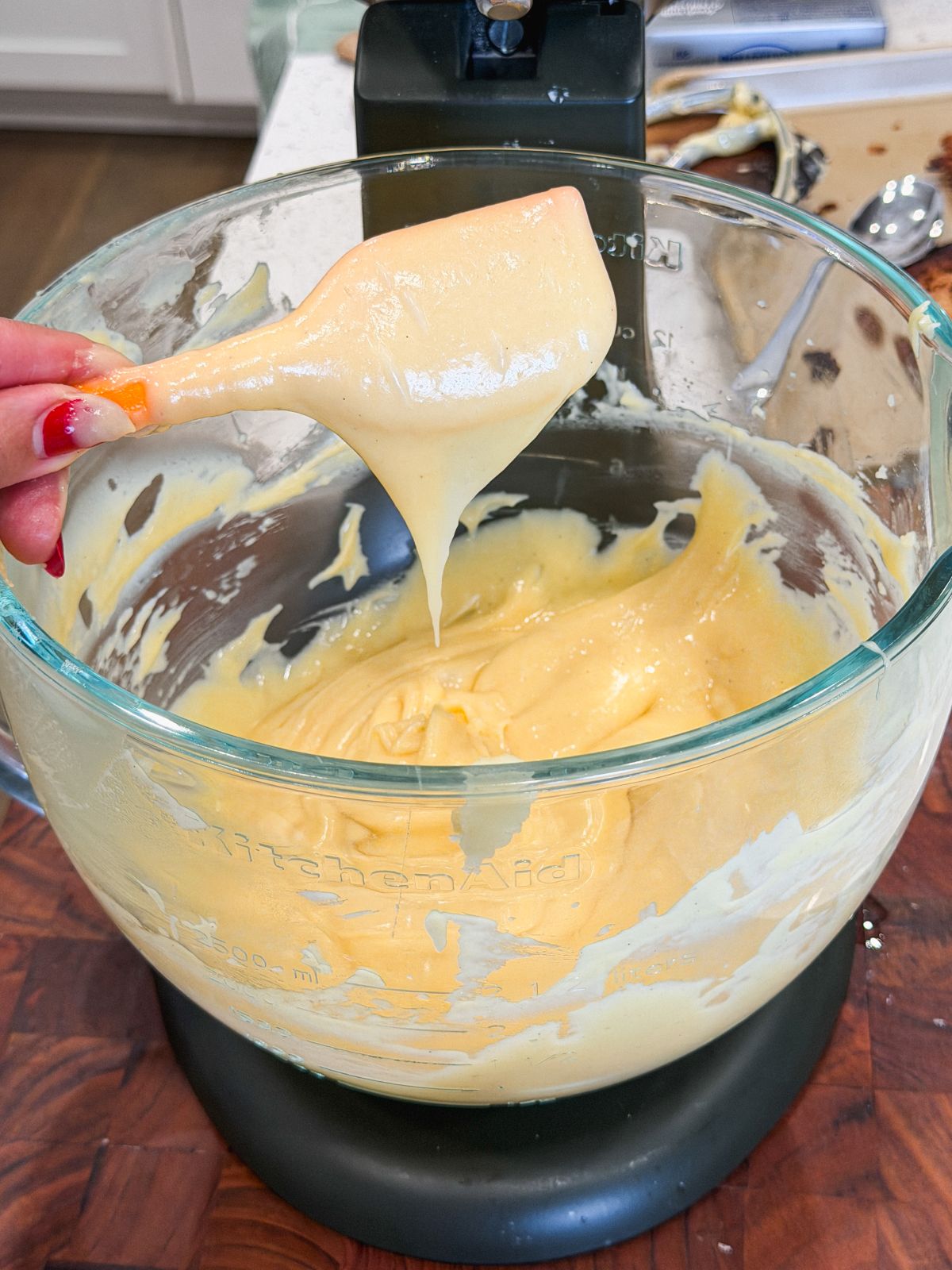 A hand holding a spatula with choux pastry over a glass mixing bowl attached to a stand mixer on a wooden countertop. The creamy batter drips from the spatula back into the bowl. The choux pastry has fallen off the spatula leaving behind a smooth, tapered V shape