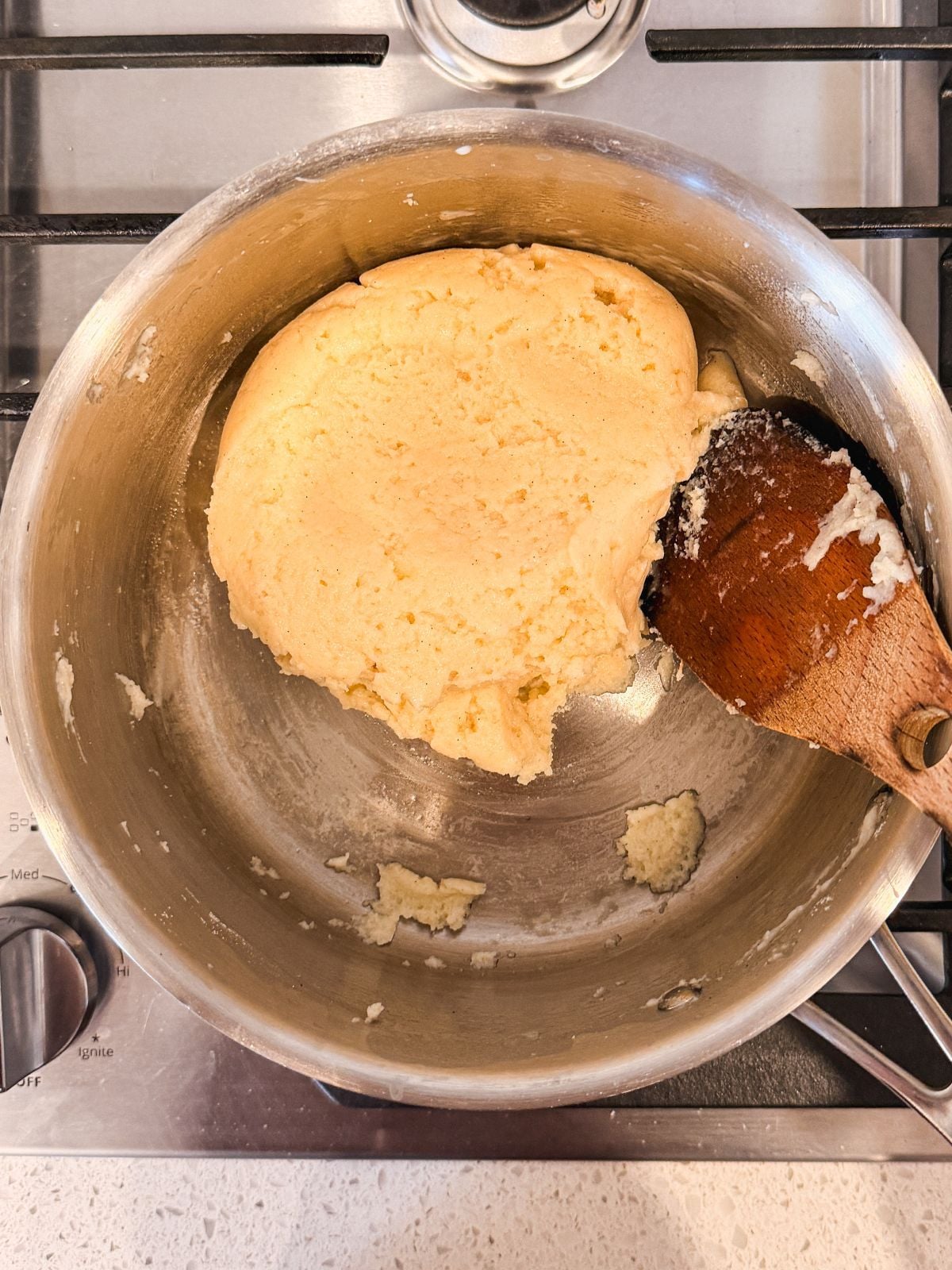 A ball of choux pastry dough sits in a metal saucepan on a stove, with a wooden spoon resting beside it.