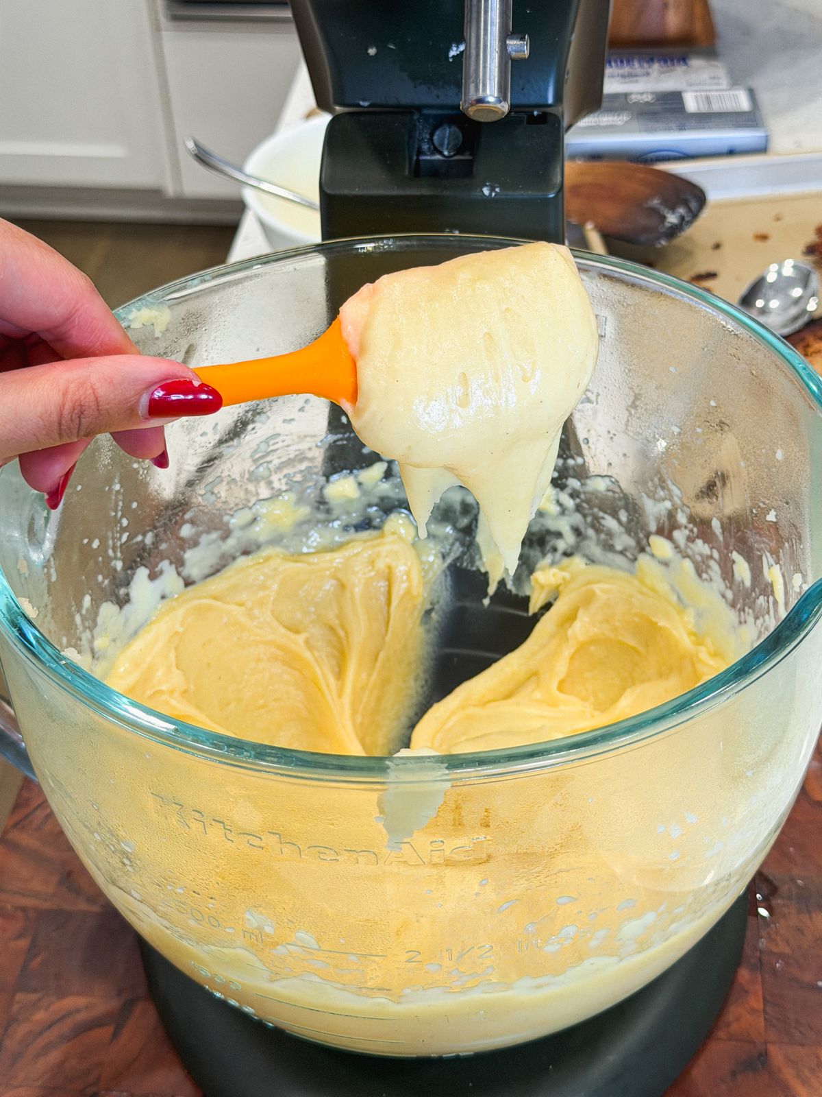 A hand holding an orange spatula with thick, choux pastry above a glass mixing bowl attached to an electric stand mixer on a kitchen counter.Pastry has fallen off leaving a craggly shape behind